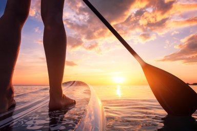 Stand up paddle boarding or standup paddleboarding on quiet sea at sunset with beautiful colors during warm summer beach vacation holiday, active woman, close-up of water surface, legs and board