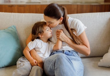 Loving Mom Kissing And Embracing Little Daughter While Sitting Together On Couch